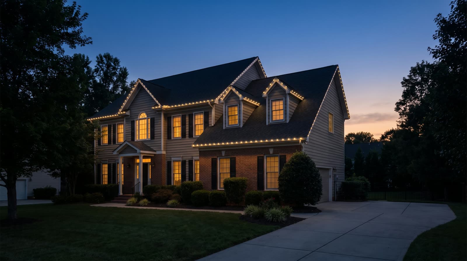 Two-story tan siding and brick house at blue hour, warm white roofline dots tracing every gable, dormer and eave above a lit front portico