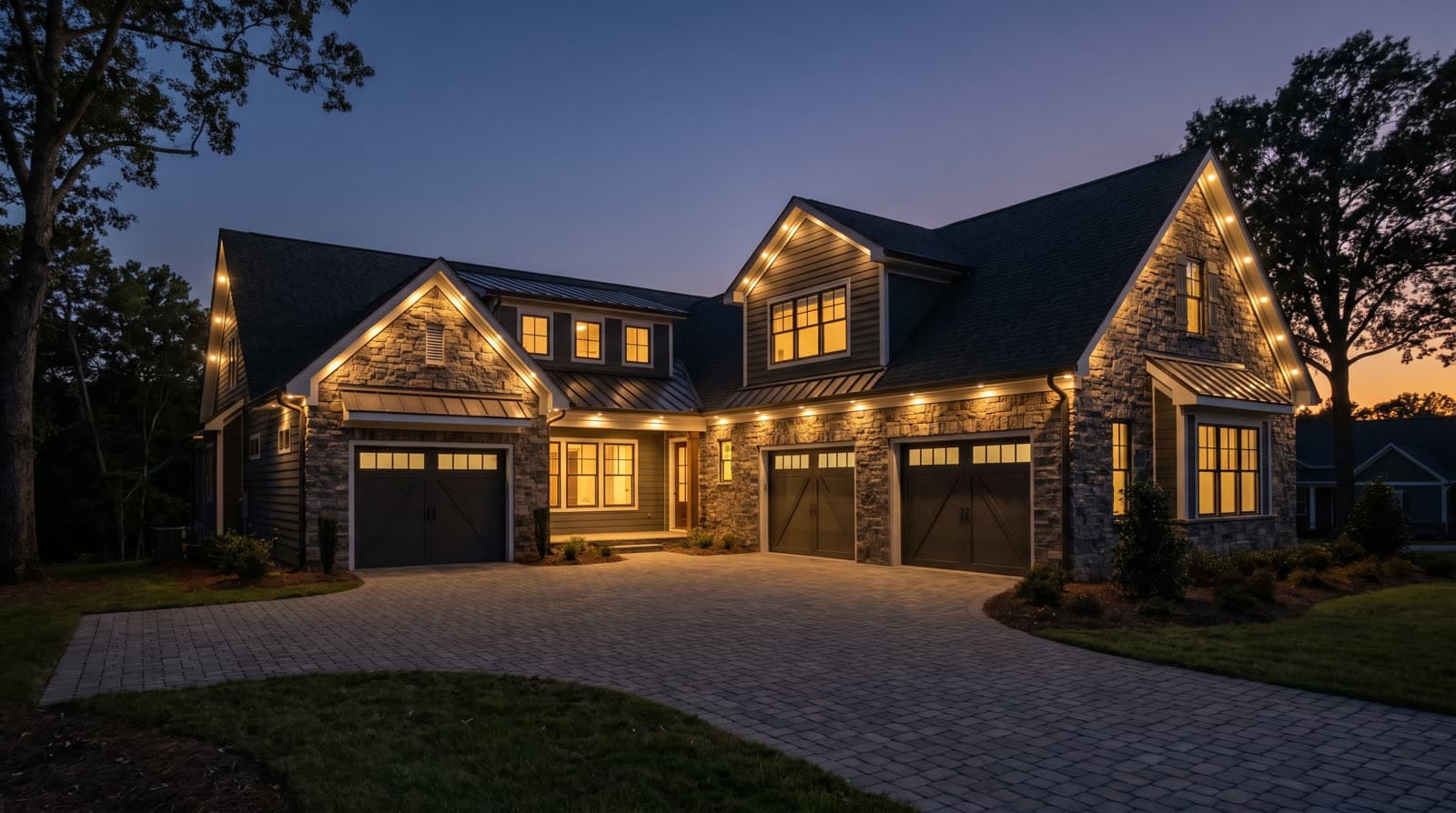 Large two-story stone and siding house at blue hour with warm white lighting tracing every roofline and gable, three-car garage