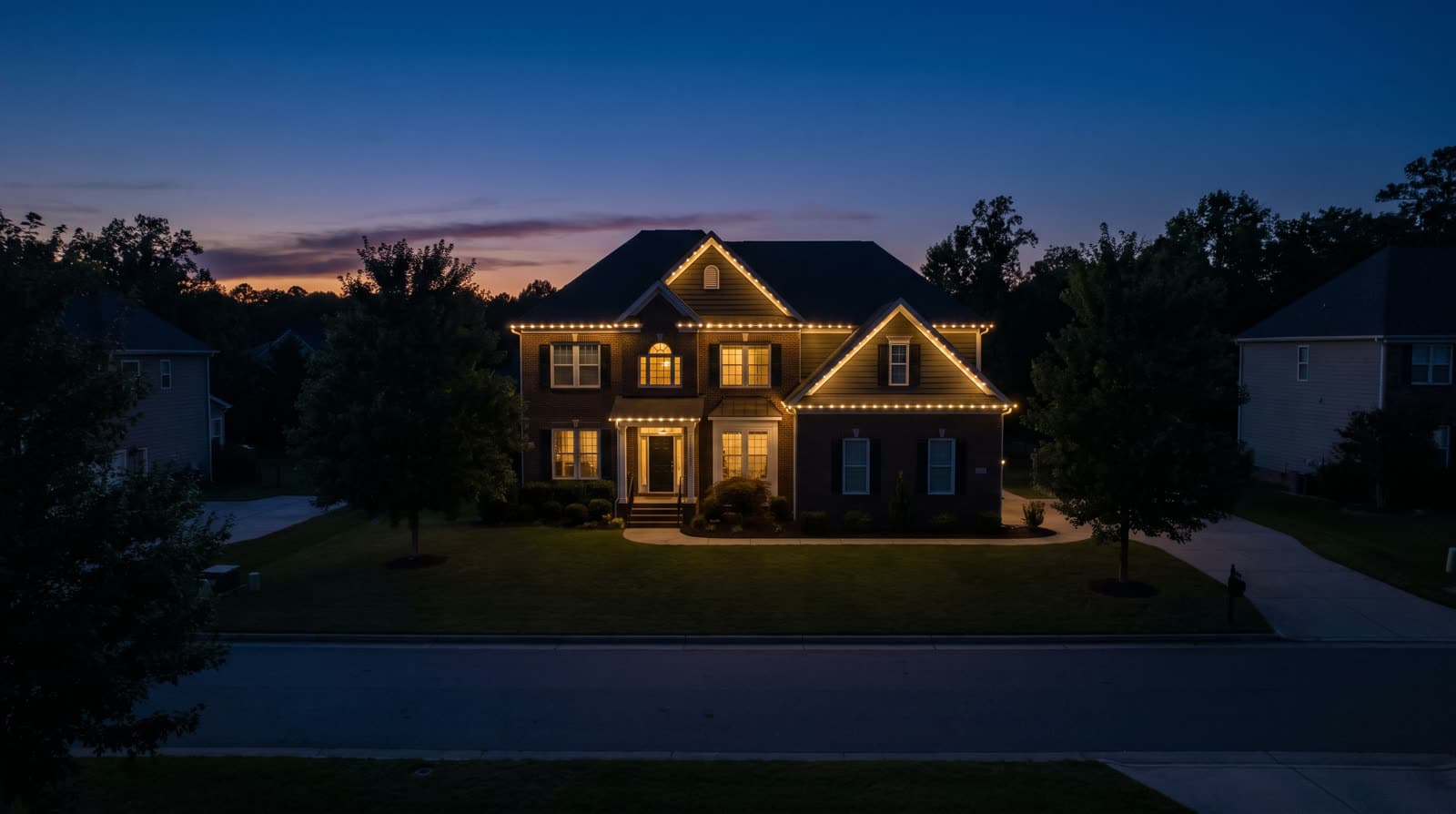 Two-story brick house at twilight seen from across the street, warm white roofline lighting tracing every gable, tall trees flanking the lot