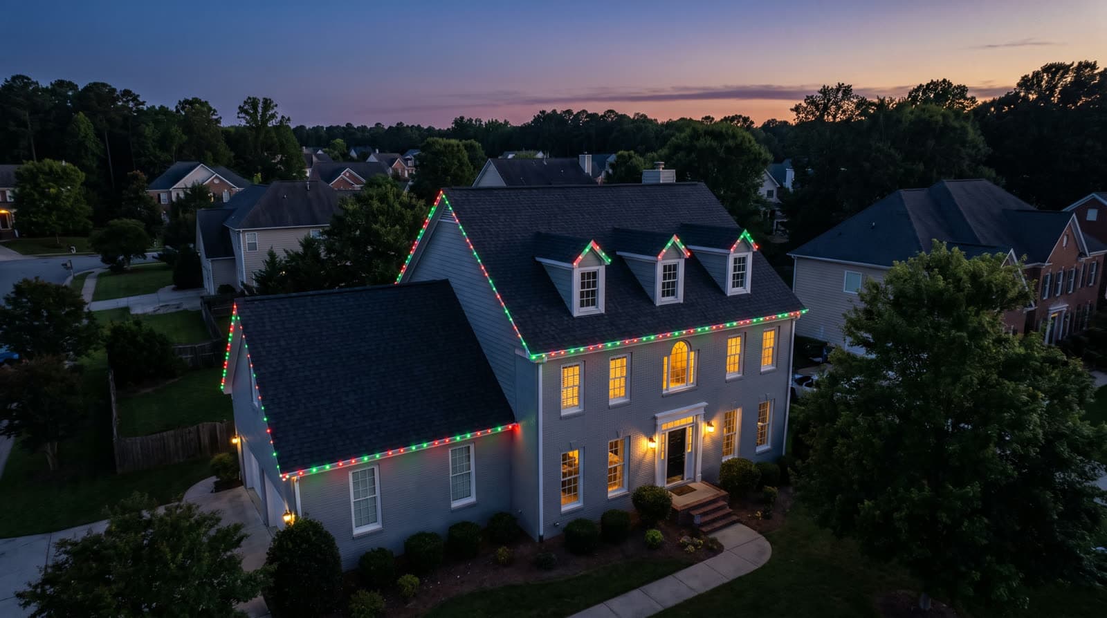 Aerial view of a two-story colonial house at sunset, red and green lights tracing every roofline gable, illuminated windows below a fading orange and purple sky