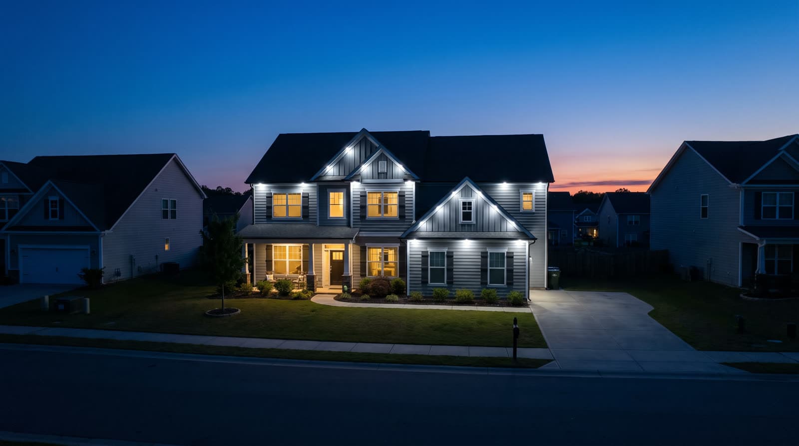 Two-story pale grey house with a front gable and a single dormer, photographed straight on from the street at deep blue hour with an orange sunset band low on the horizon, crisp warm white roofline lights tracing every eave, gable and dormer, warm amber windows, neighboring houses close on both sides