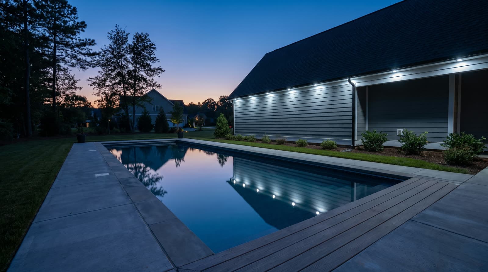 Long gray-sided house with a steep dark gable roof and a covered porch wing on white posts, seen from a rectangular pool's corner at blue hour, small bright white lights running the entire eave line, a deep violet sky behind silhouetted pines, wood decking in the foreground.