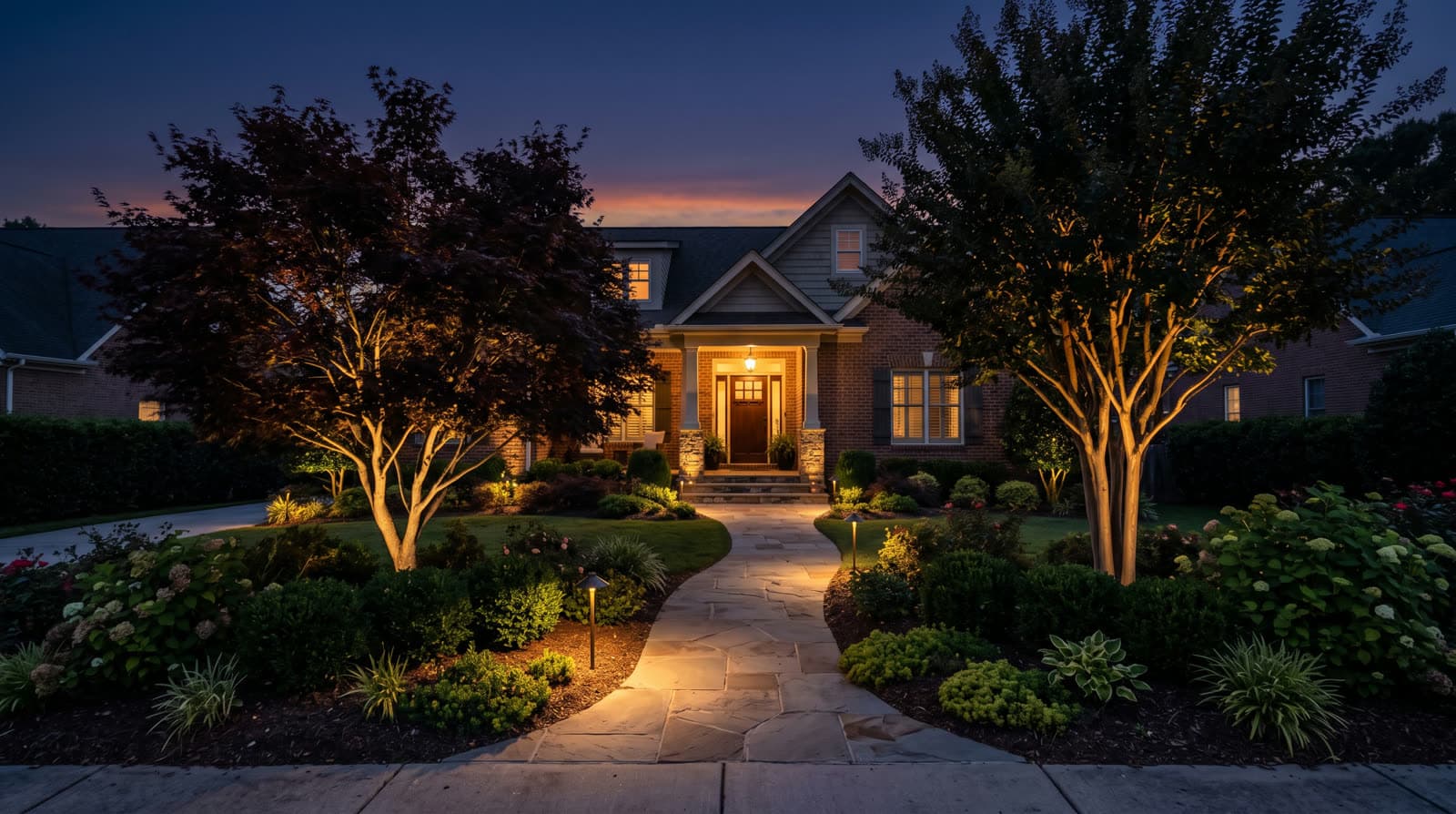 Brick house front entry at twilight framed by two ornamental trees, with warm white landscape path lighting along a curved stone walkway